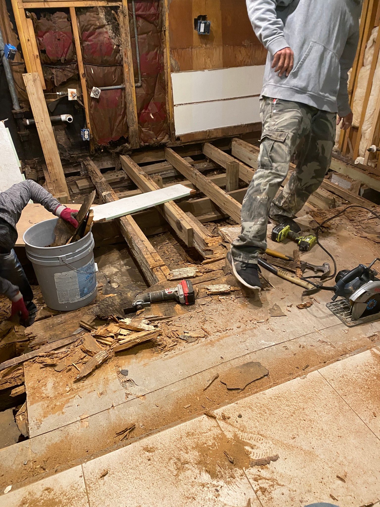 Home renovation scene with exposed floor joists, tools, and a worker standing in a gutted room