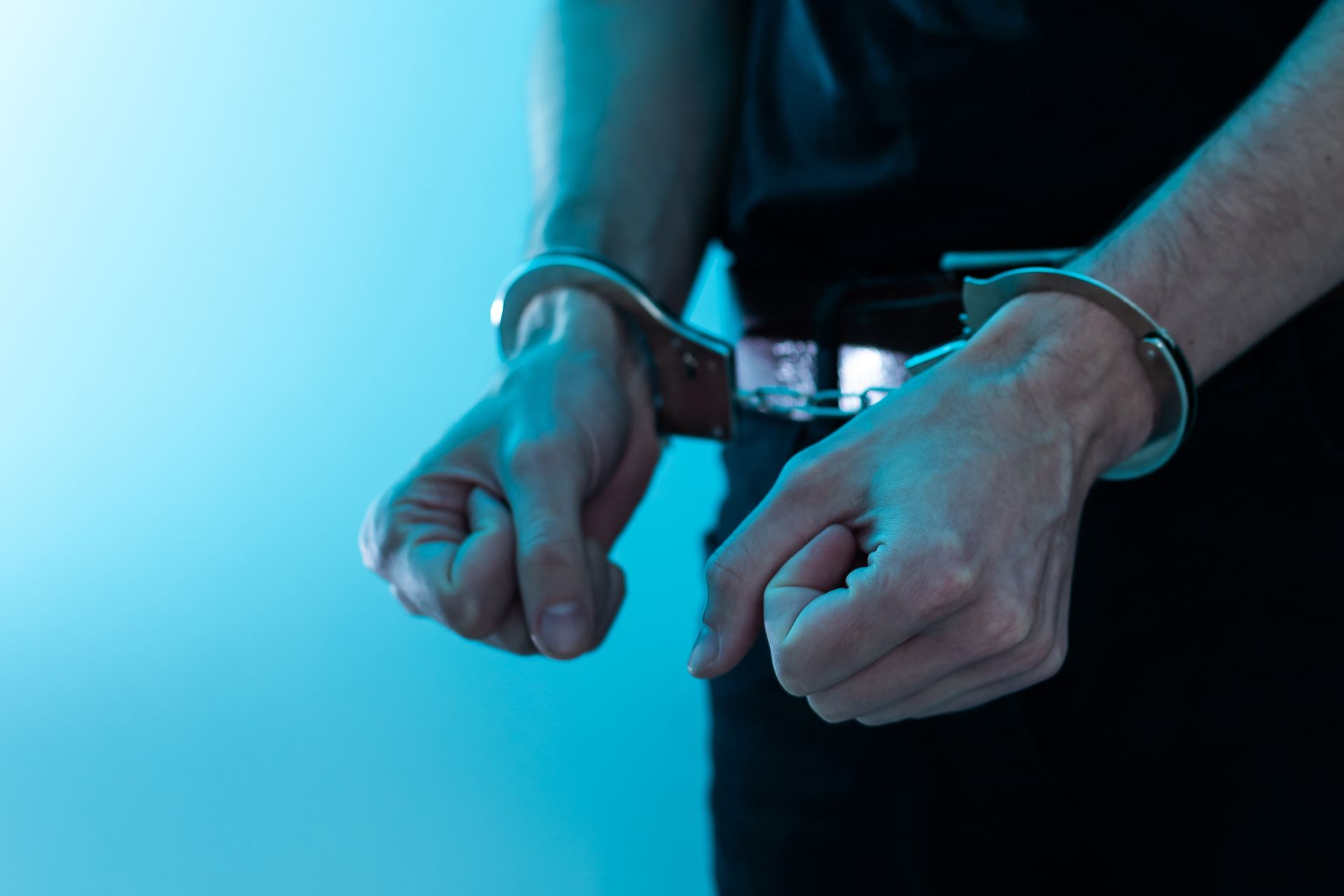 Person's hands in handcuffs against a light blue background, suggesting arrest.