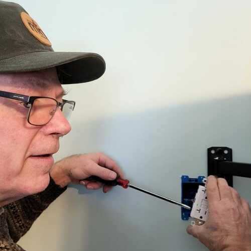 A man wearing glasses and a hat is working on a wall with a screwdriver