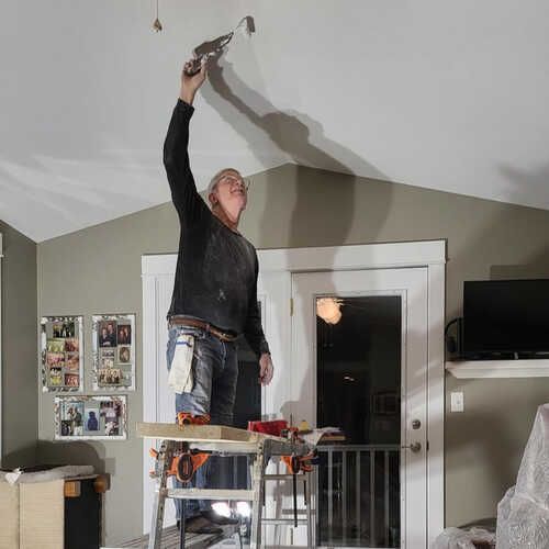 A man is standing on a ladder in a living room holding a spatula