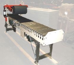 Conveyor belt system with rollers, black belt, and red arm, on metal legs and frame, in a factory setting.