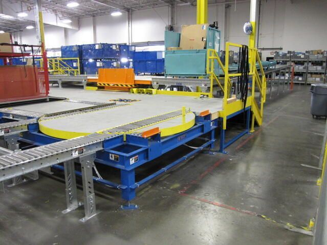 Automated conveyor system with rotating platform in a warehouse; blue, yellow, and gray.