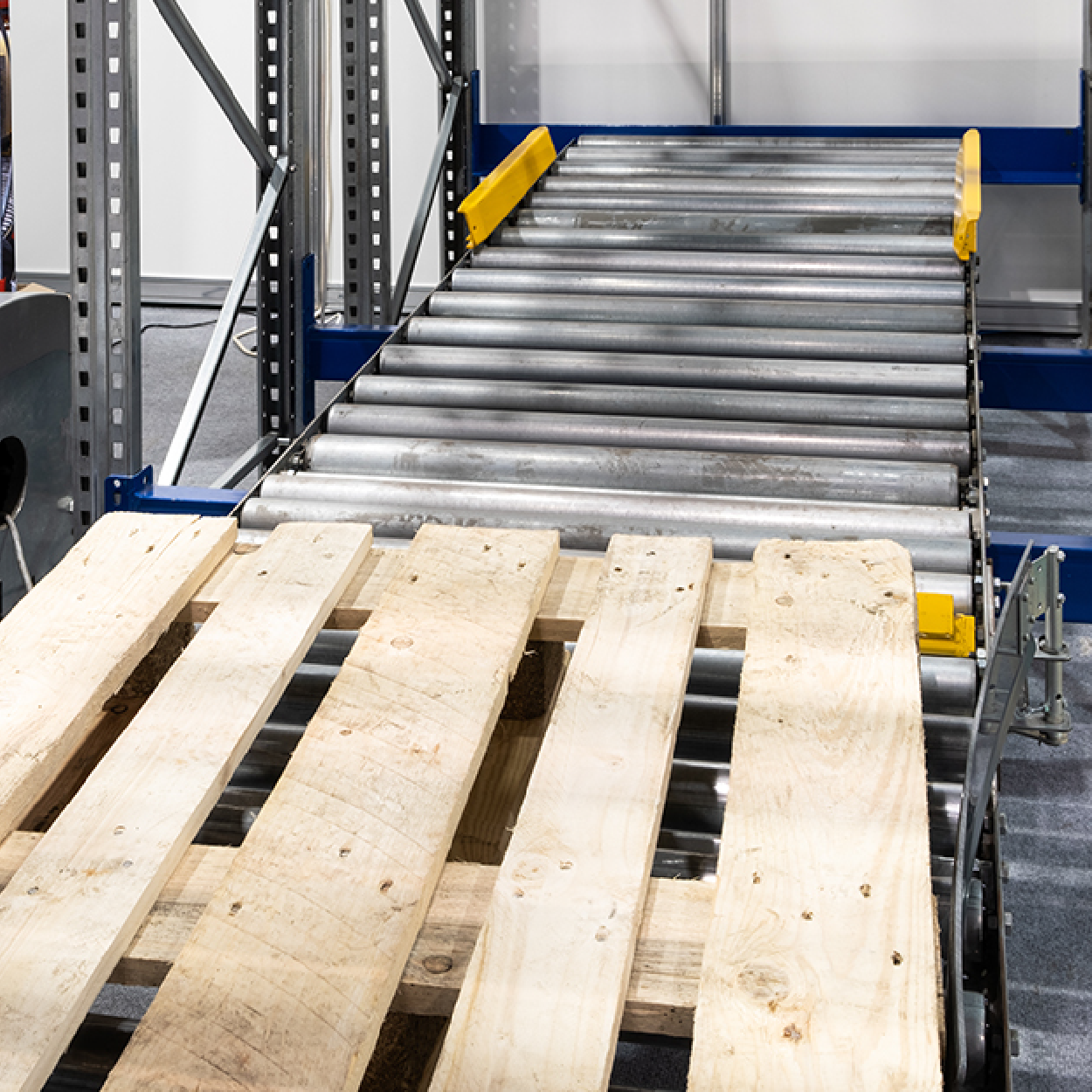 A wooden shipping pallet sits at the start of a metal roller conveyor track inside an industrial warehouse rack system.