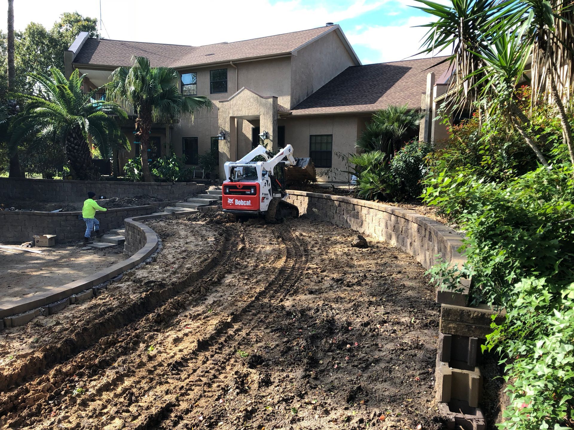 Construction site with a Bobcat excavator on muddy driveway near a house. A worker is present.