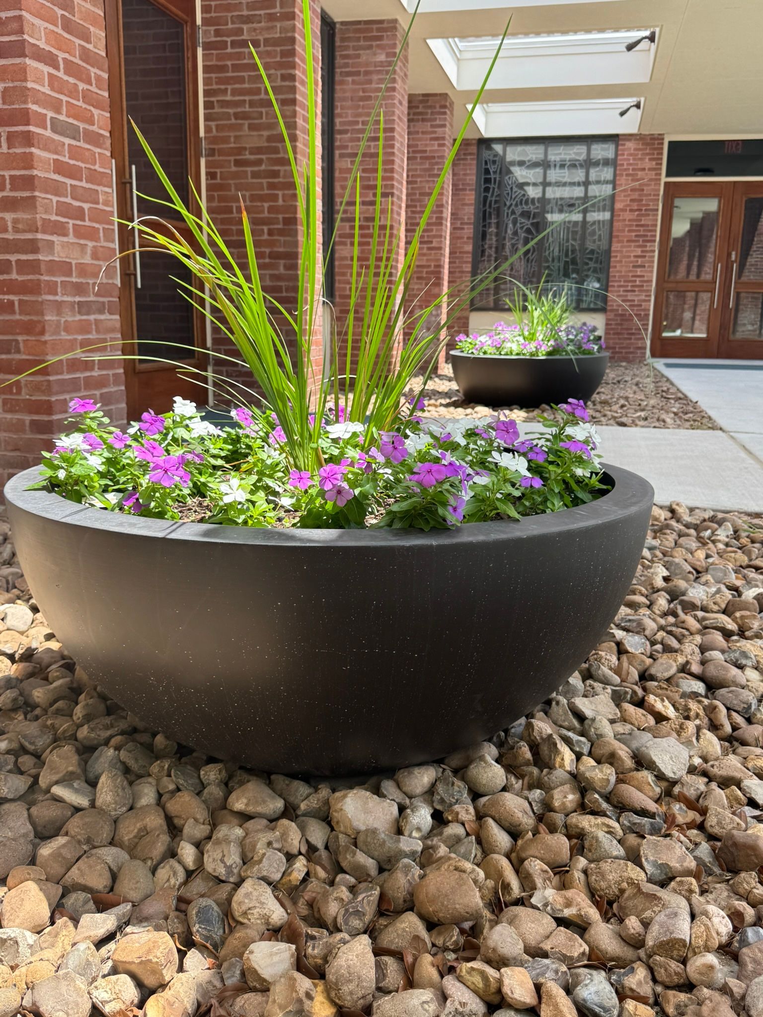 A large black planter filled with purple and white flowers in front of a brick building.