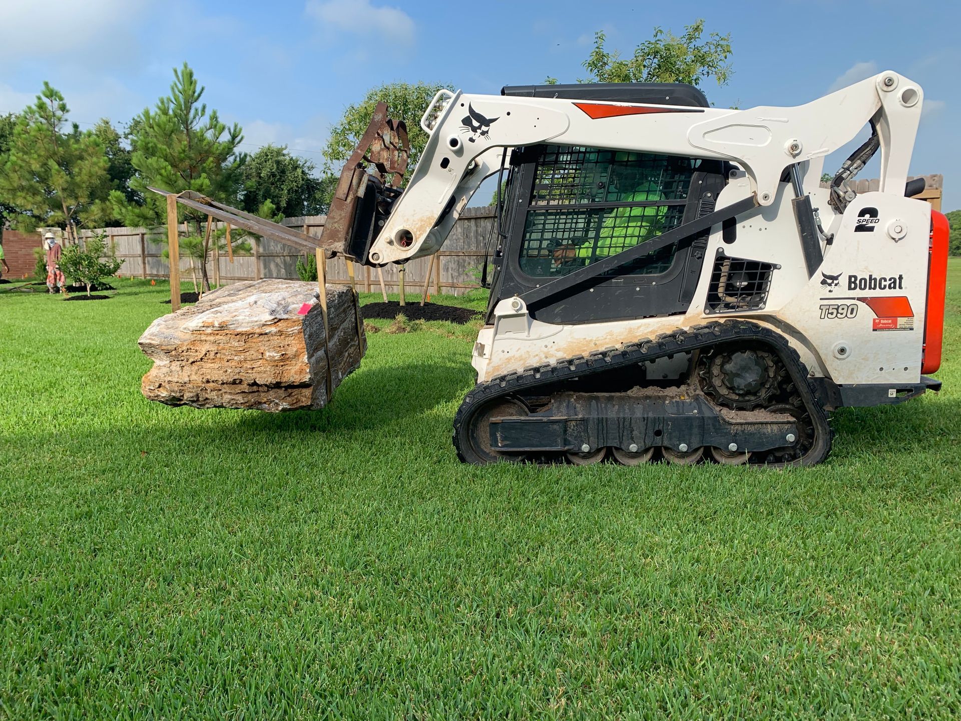 A bobcat is moving a large rock in a lush green field.