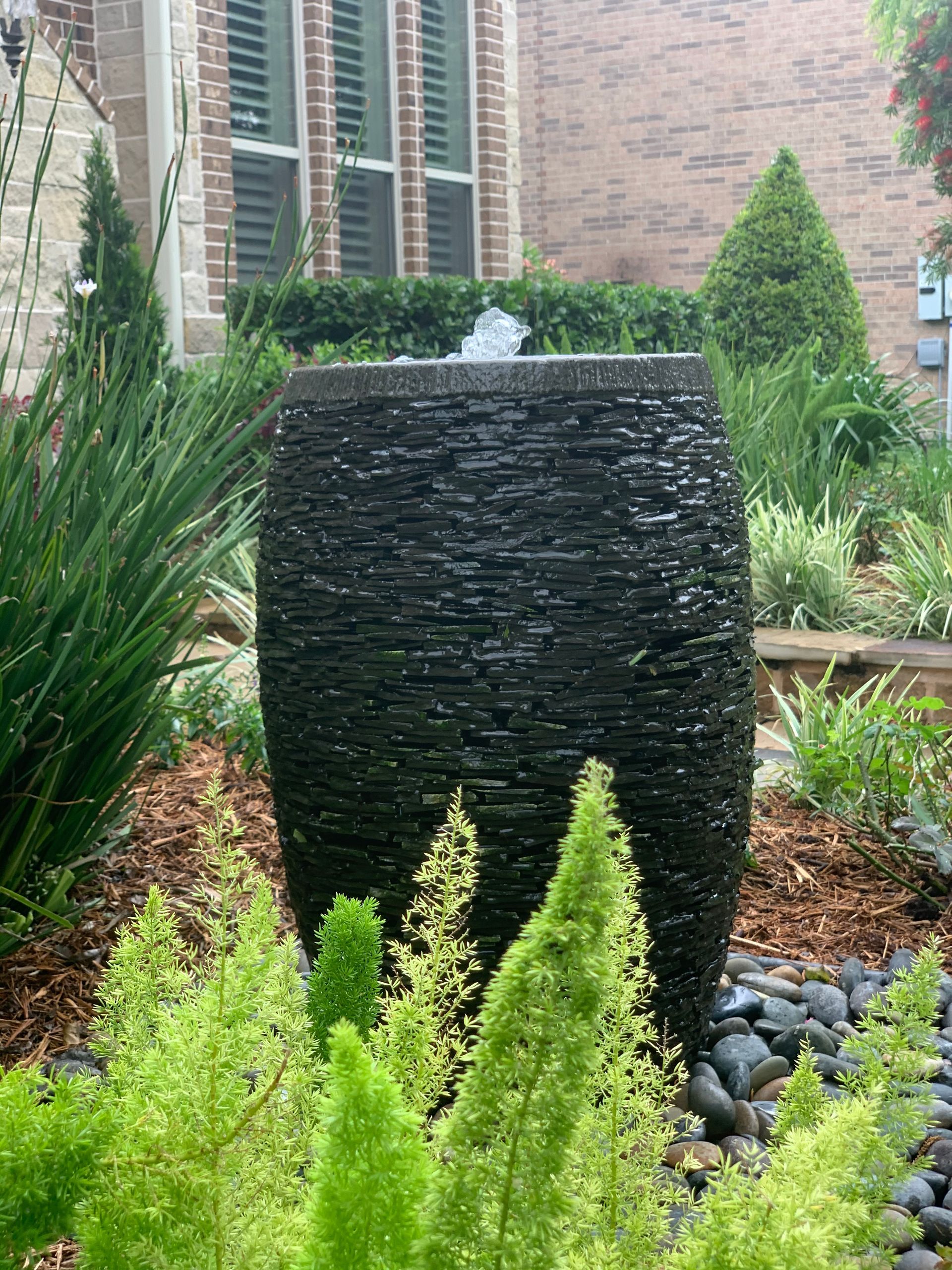 A fountain in a garden with a brick building in the background.