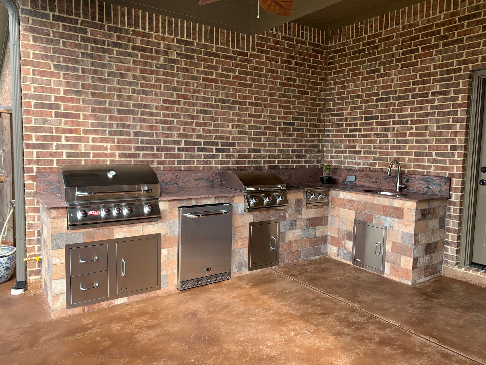 A large outdoor kitchen with a brick wall and stainless steel appliances.