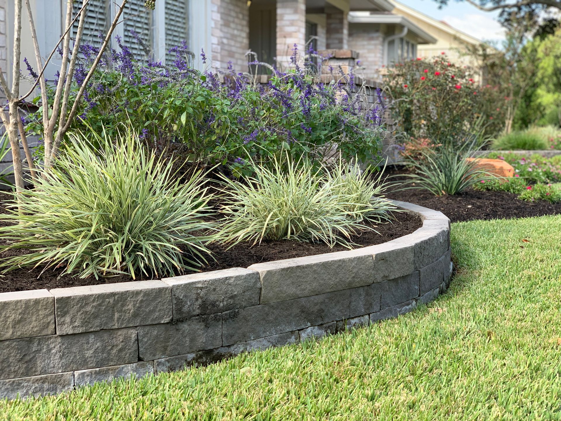 A lush green lawn with a stone wall and flowers in front of a house.
