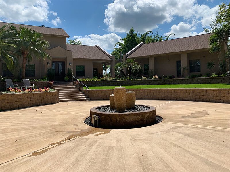 A large beige house with a fountain in the driveway and manicured landscaping under a blue sky.