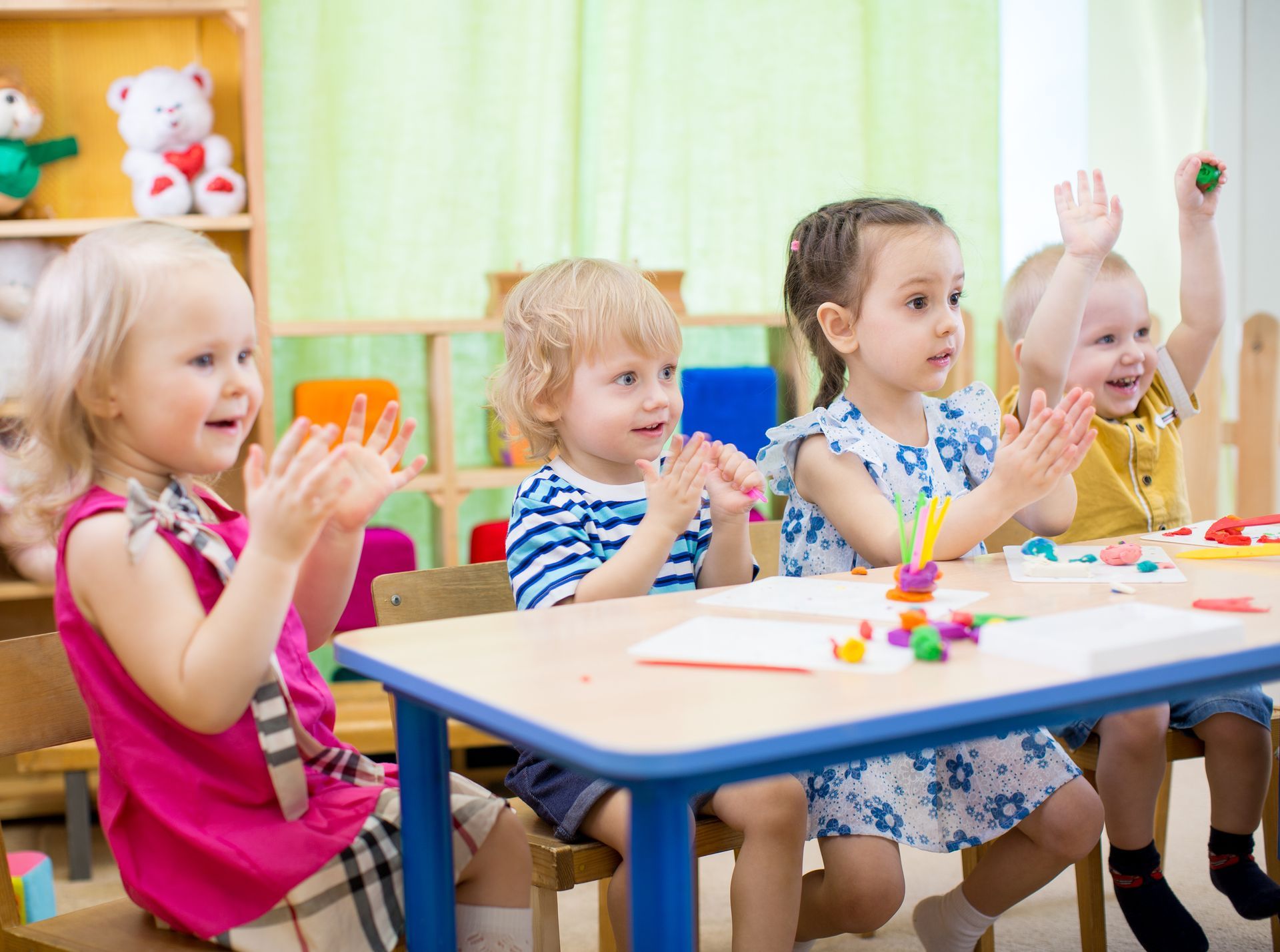 a group of children sit at a table with their hands up