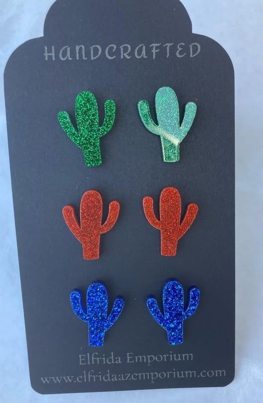 Cactus-shaped glitter earrings in green, blue, and red on a gray display card.