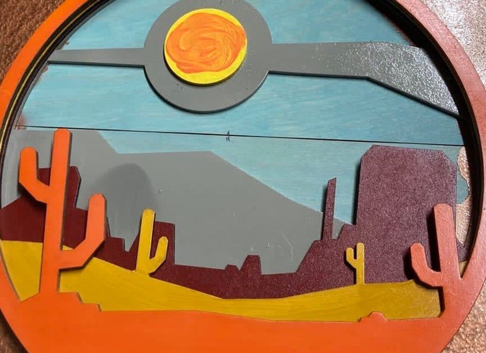 Wooden desert landscape art with a sun, cacti, and layered mountains in shades of orange, yellow, gray, and blue.