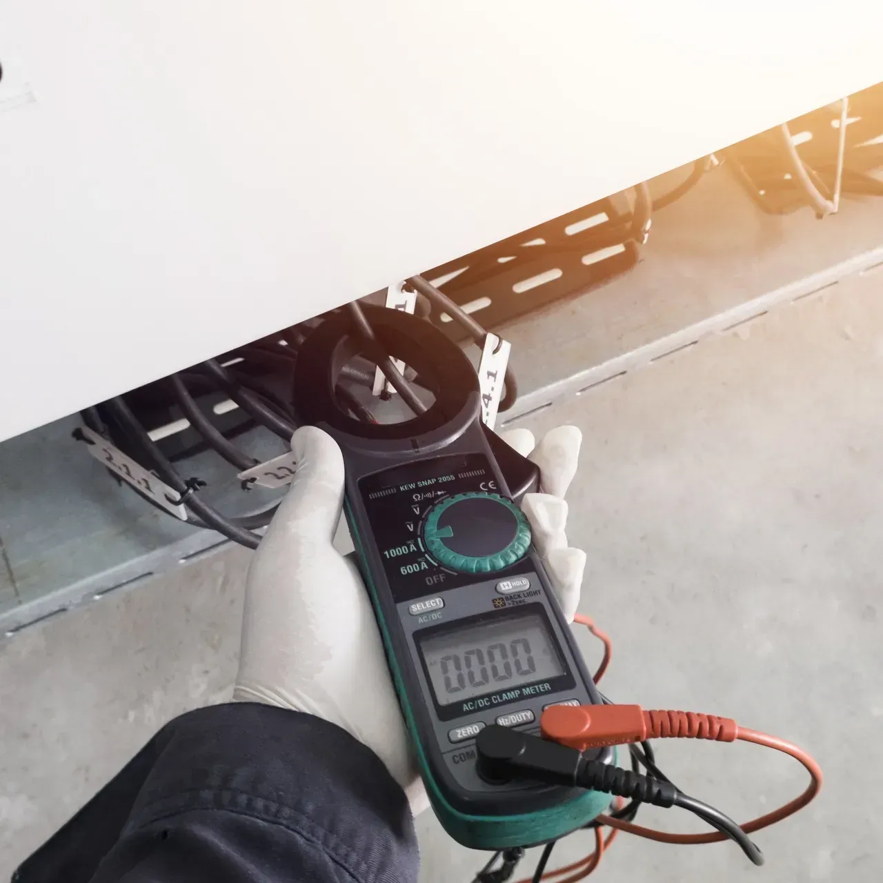 A person in gloves uses a digital multimeter to measure current on a wire, indoor setting.