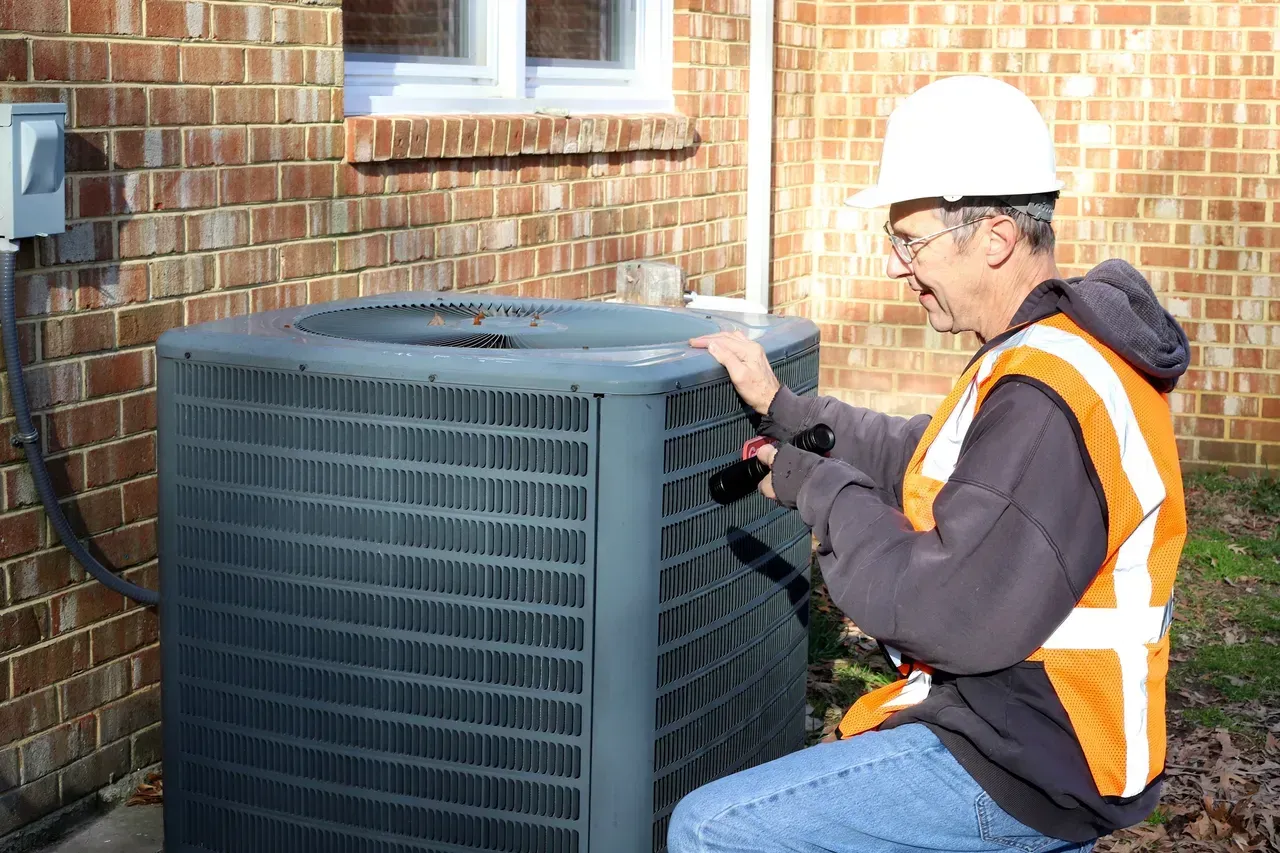 HVAC technician inspecting an air conditioning unit outside a brick building. Wearing a hard hat and safety vest.