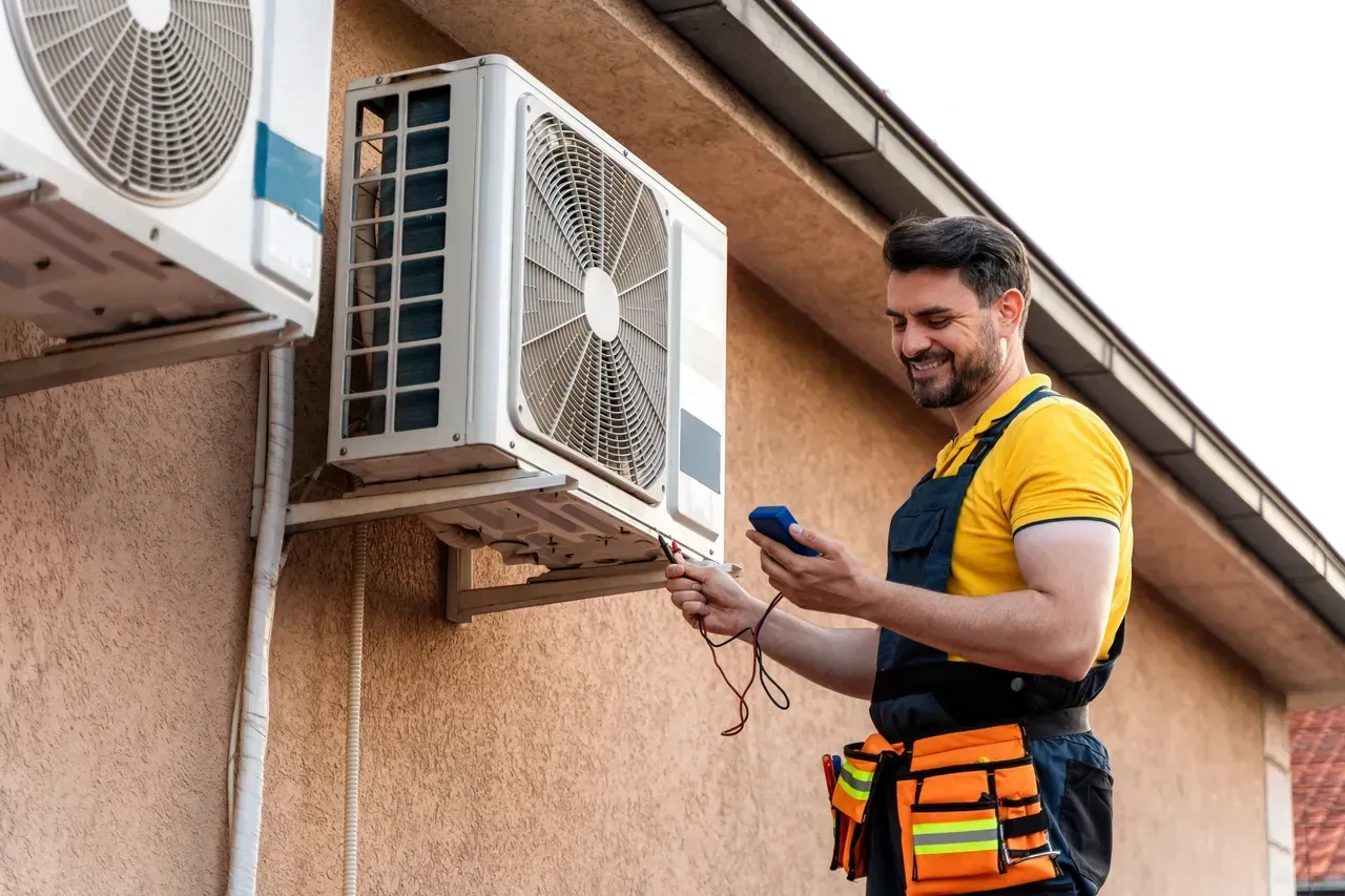 HVAC technician examining an outdoor air conditioning unit with a device, smiling, in front of a tan wall.
