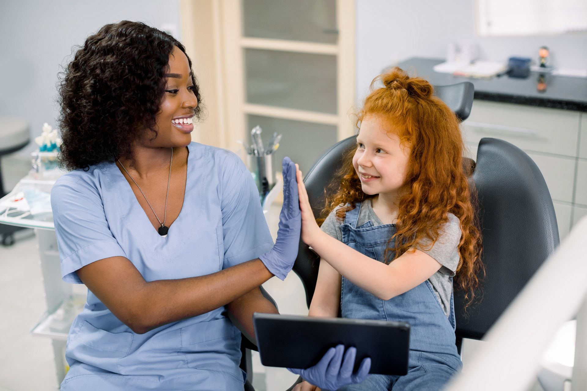 A dentist and a child are sharing a high five in a clinic with a tablet and dental tools nearby