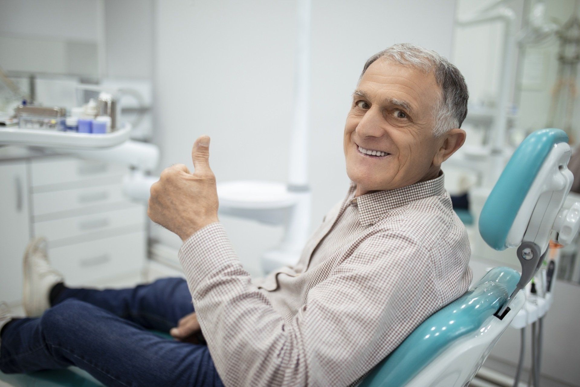 An elderly man is sitting in a dental chair and giving a thumbs up.