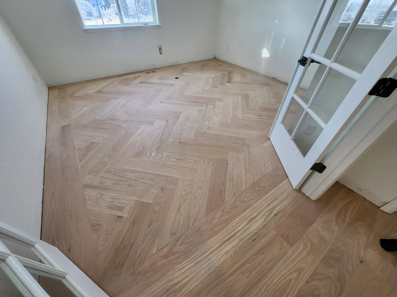 Herringbone wood floor in a room with white walls, a window, and a white door.