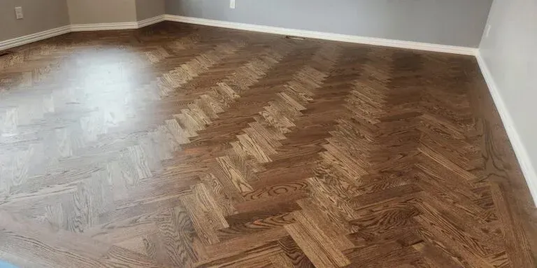Herringbone pattern wood flooring in a room with white trim and light gray walls.