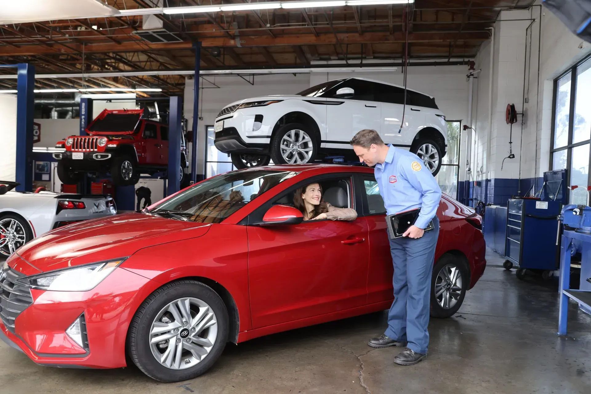 A mechanic talking with a driver in a red car at a repair shop with other vehicles on lifts.