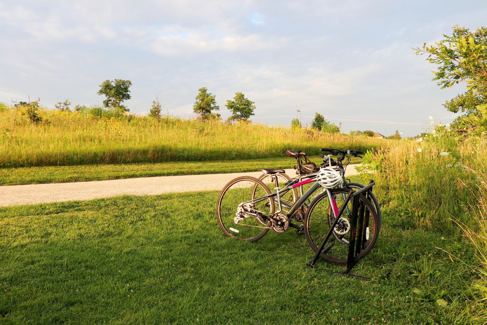 Two bicycles parked beside a gravel path in a grassy field under a partly cloudy sky.