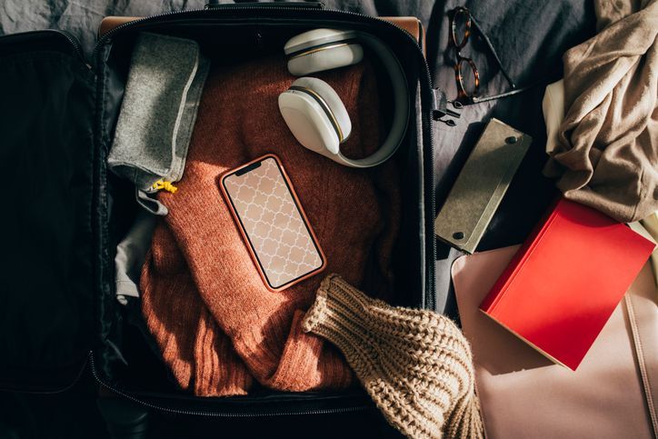 A neatly packed open suitcase sits on a bed, filled with cozy travel essentials. Inside the suitcase, a rust-colored sweater, a beige knitted garment, and a gray pouch are carefully arranged. A smartphone with a patterned case rests on top of the sweater. A pair of white over-ear headphones is also placed inside. Surrounding the suitcase are a pair of glasses, a red notebook, a beige purse, and a black portable speaker, all suggesting preparation for a trip. The scene is bathed in warm, natural light.