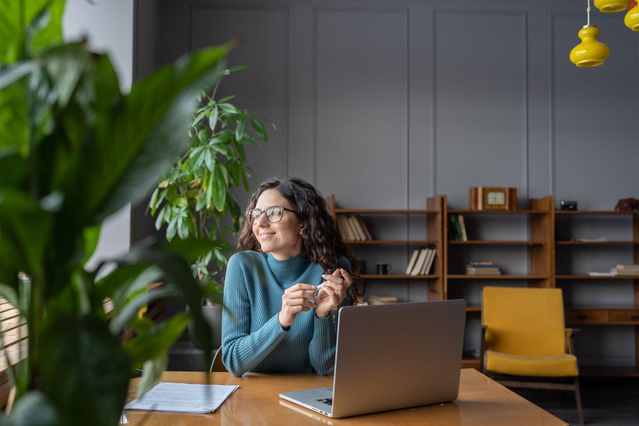 A woman with long dark hair and glasses sits at a wooden desk with a laptop, smiling thoughtfully while holding her glasses. She is in a modern, cozy workspace with large green plants, bookshelves, and warm natural light.