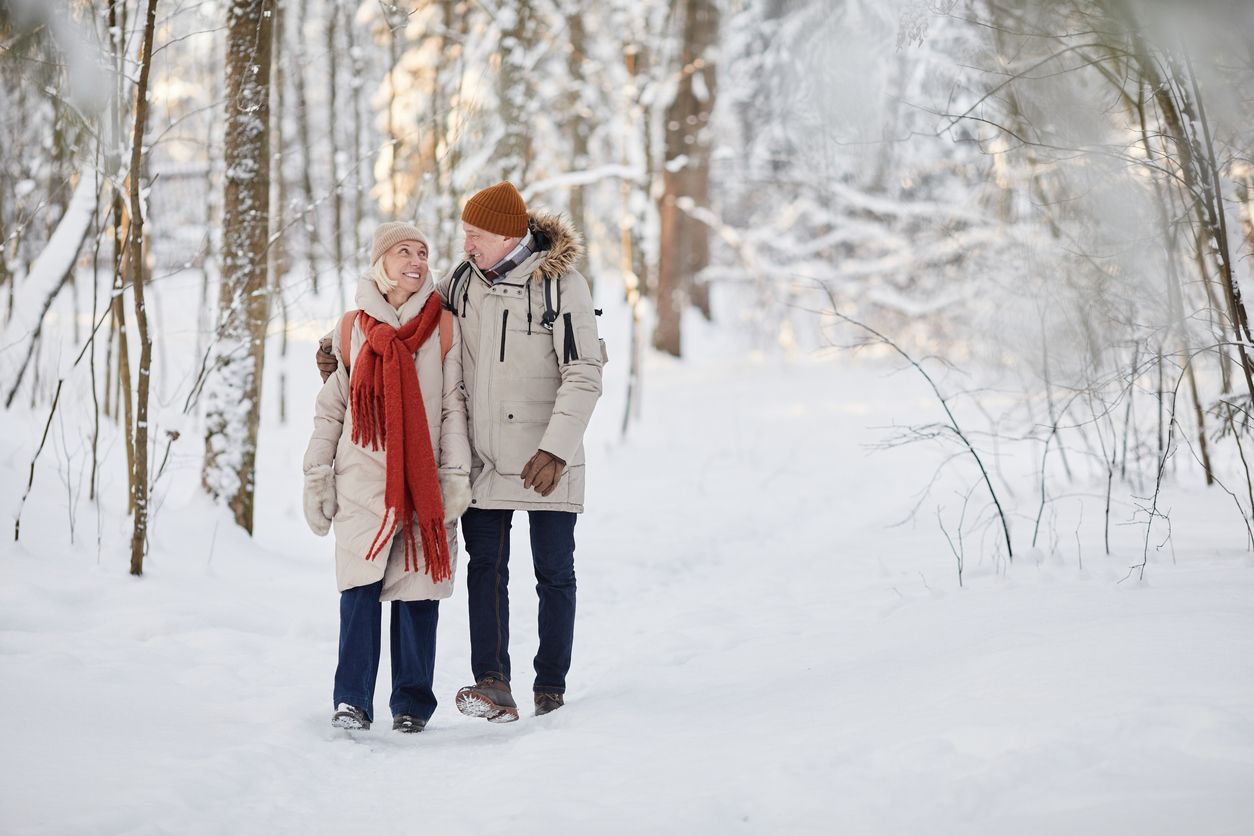 An older couple walks arm in arm through a snowy forest, dressed in warm winter coats, hats, and scarves, smiling as they enjoy a peaceful winter stroll.