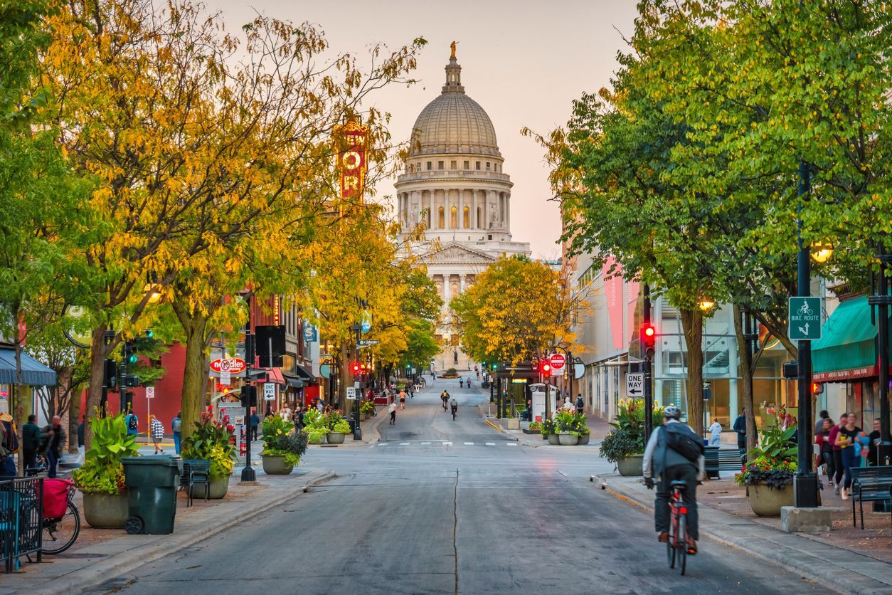 Downtown Madison street view in autumn, with colorful fall trees lining State Street and the Wisconsin State Capitol centered in the background.