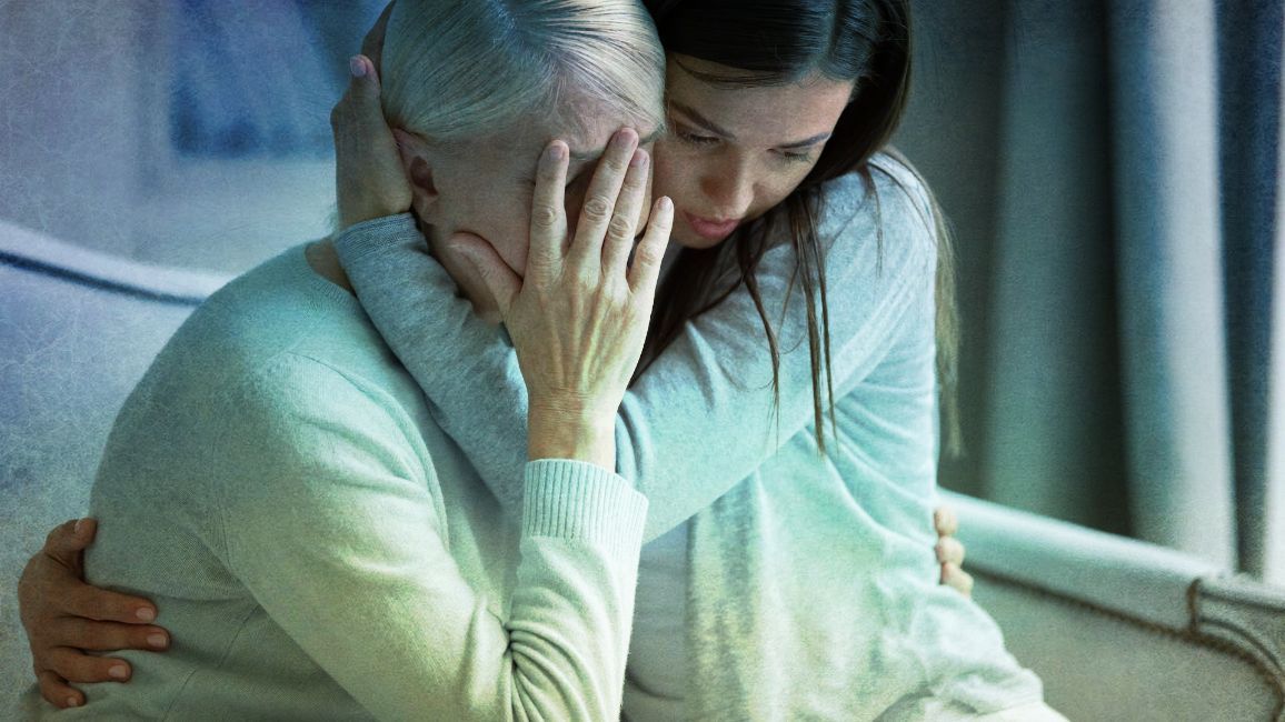 a woman is hugging an older woman who is covering her face with her hands .
