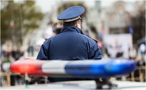 Police officer standing near a patrol car with flashing lights while monitoring a public area crowd.