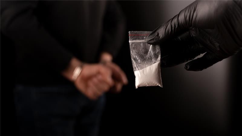 Gloved hand holding a small plastic bag of white powder as a person stands blurred in the background