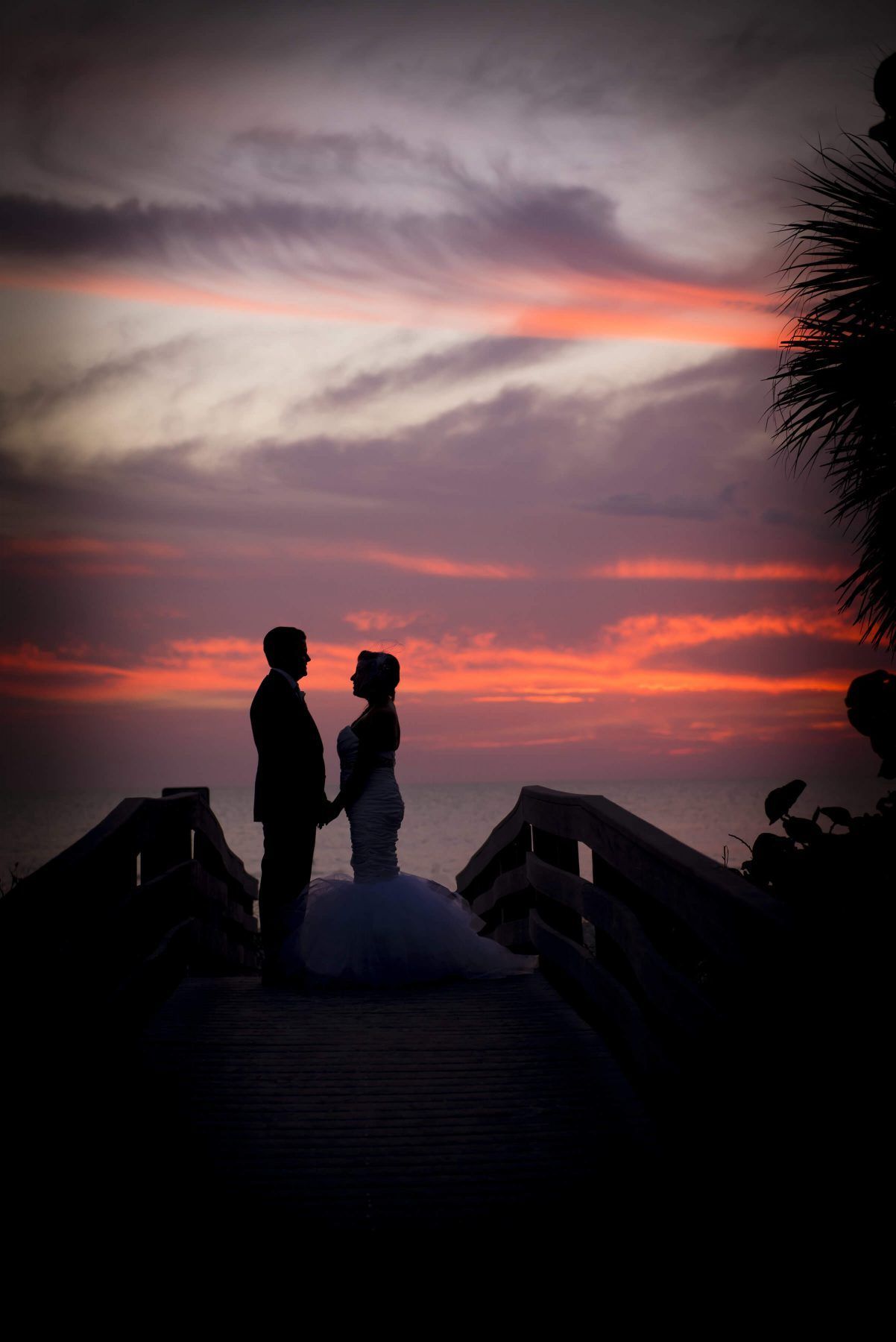 Couple silhouetted on a wooden walkway, holding hands at sunset over the ocean.