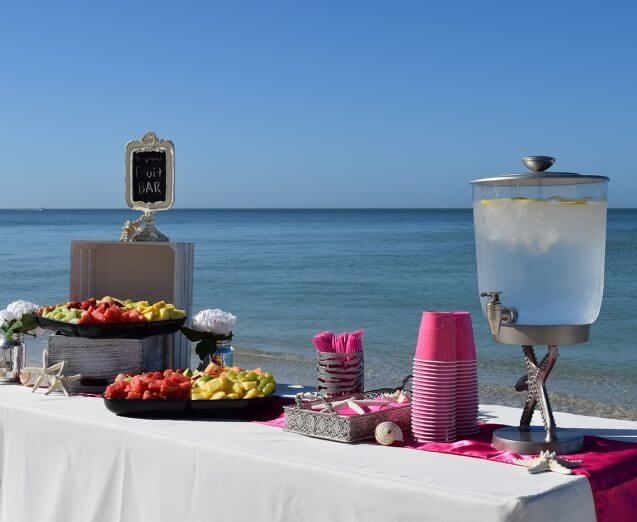 Beachside refreshment table with fruit, water dispenser, pink cups, and a sign.