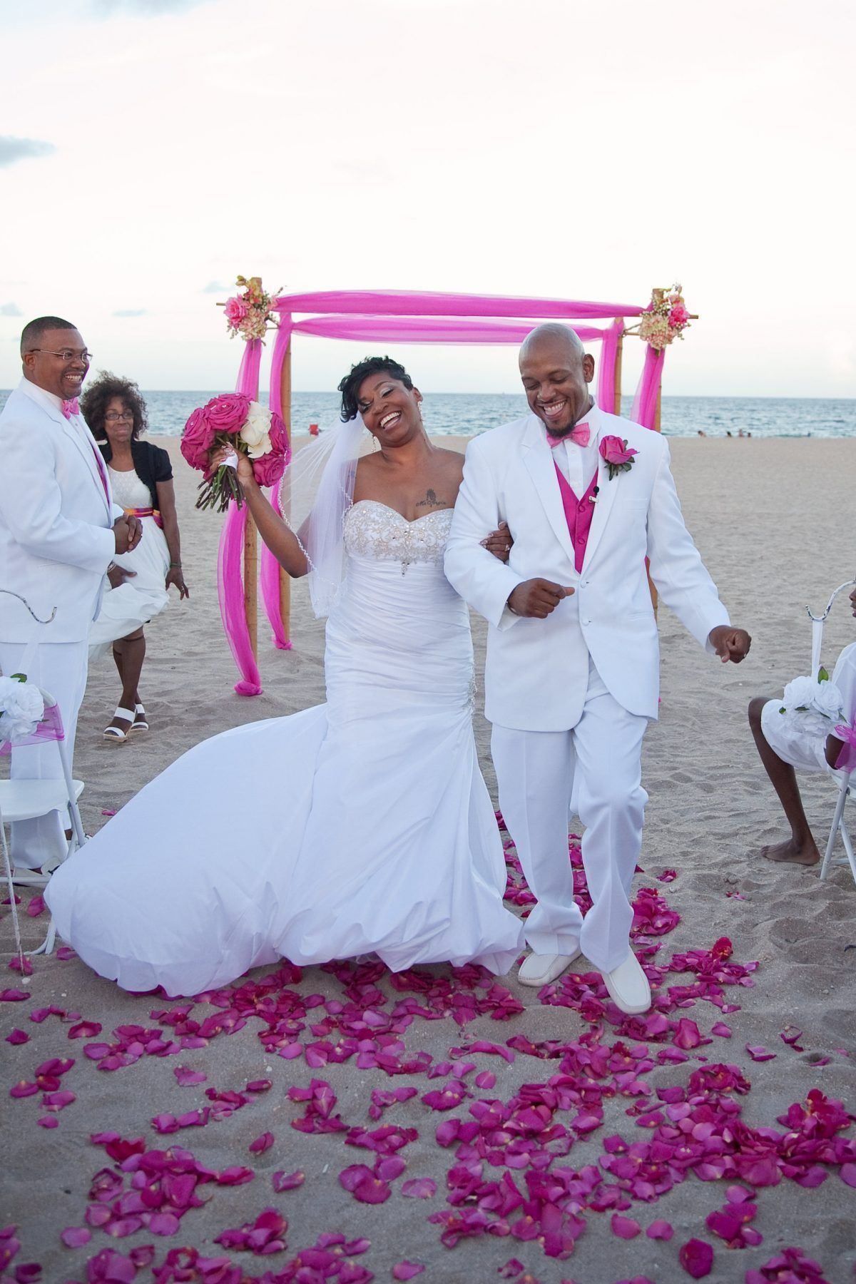 Newlyweds walk on a beach, smiling. Pink flower petals and archway. Groom in white suit, pink accents. Bride in white dress.