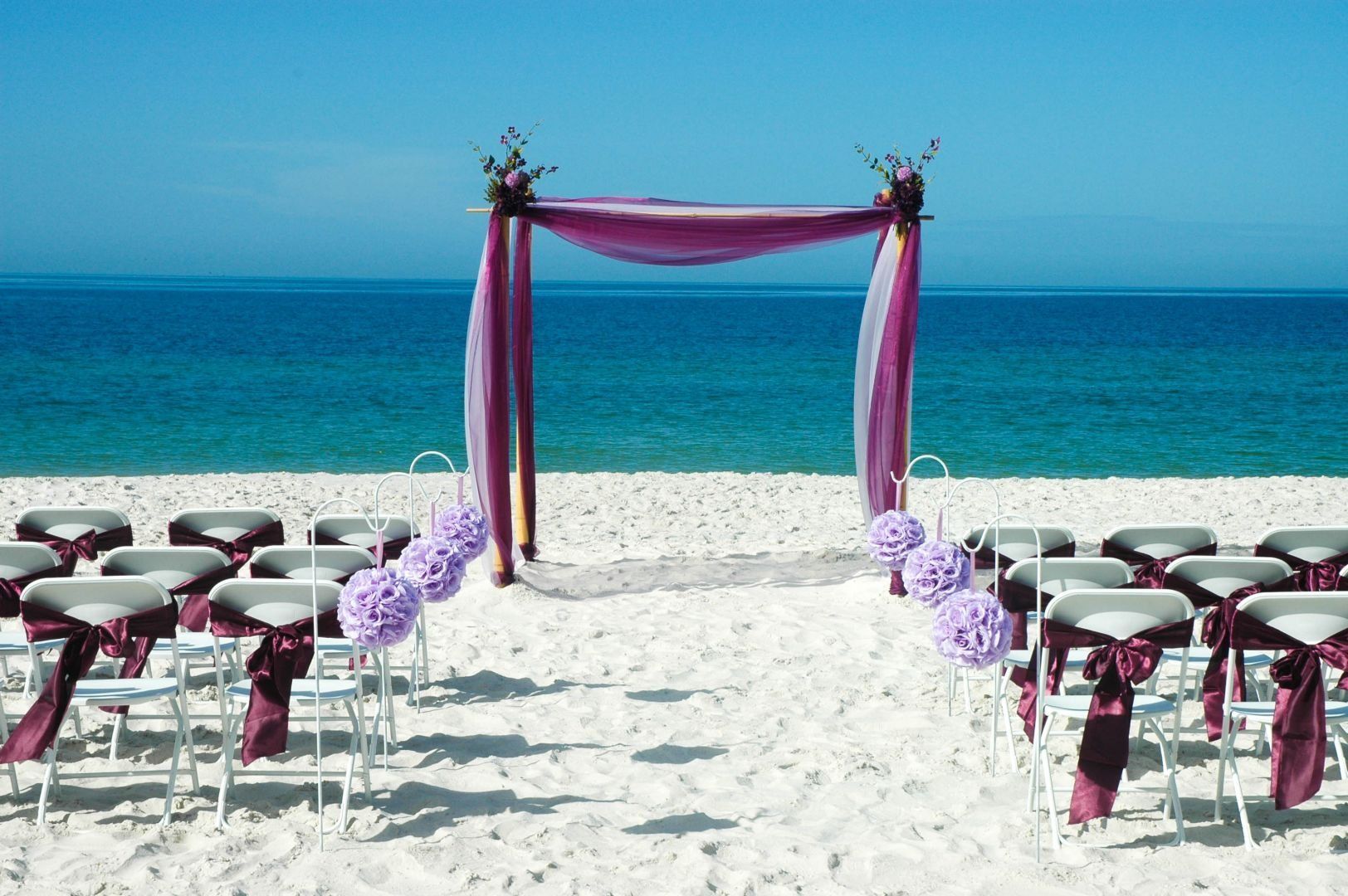 Beach wedding ceremony setup: purple arch, chairs with purple sashes, white sand, and ocean backdrop.