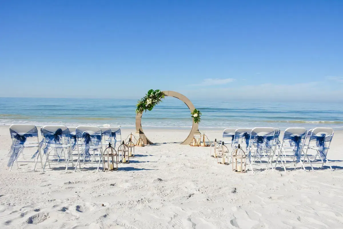 Wedding ceremony setup on a white sandy beach with ocean in the background, blue sky.