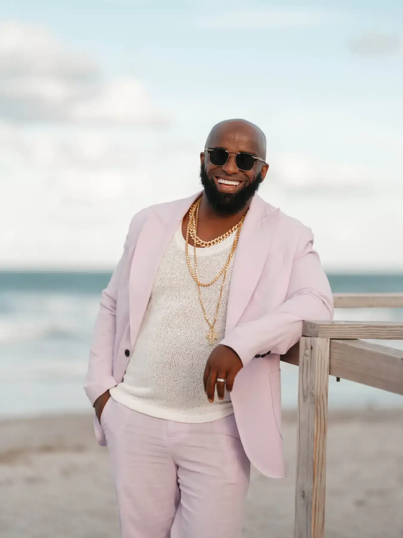 Man in light pink suit and sunglasses smiles at the beach, leaning on a wooden railing.