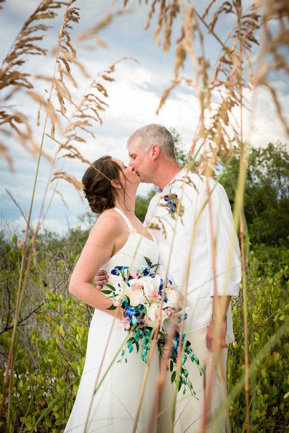 Couple kissing outdoors, framed by tall grass. The woman wears a white dress, holding a bouquet.