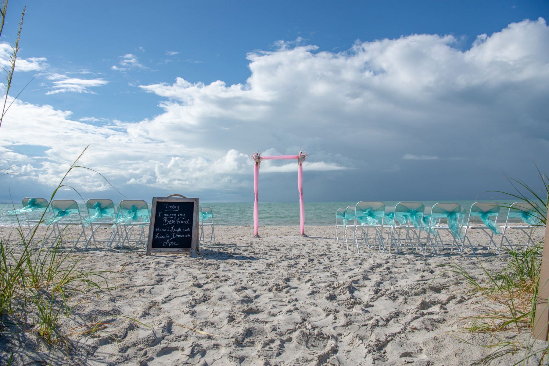 Beach wedding setup with turquoise chairs, a pink arch, and a chalkboard sign.