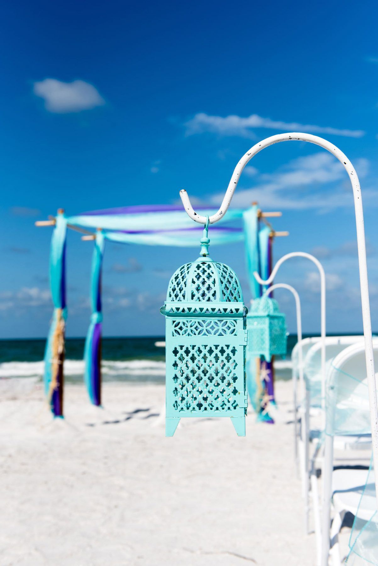 Teal lantern hanging on a white hook on a beach, wedding arch in the background, blue sky.