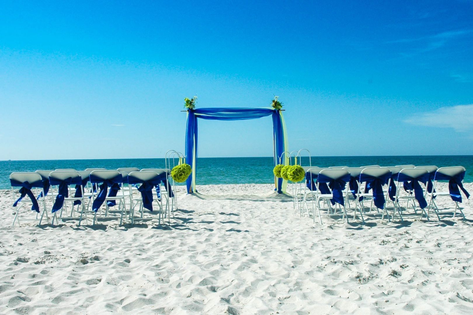 Beach wedding ceremony setup: blue archway, rows of chairs with blue sashes, on white sand, blue sky and ocean backdrop.