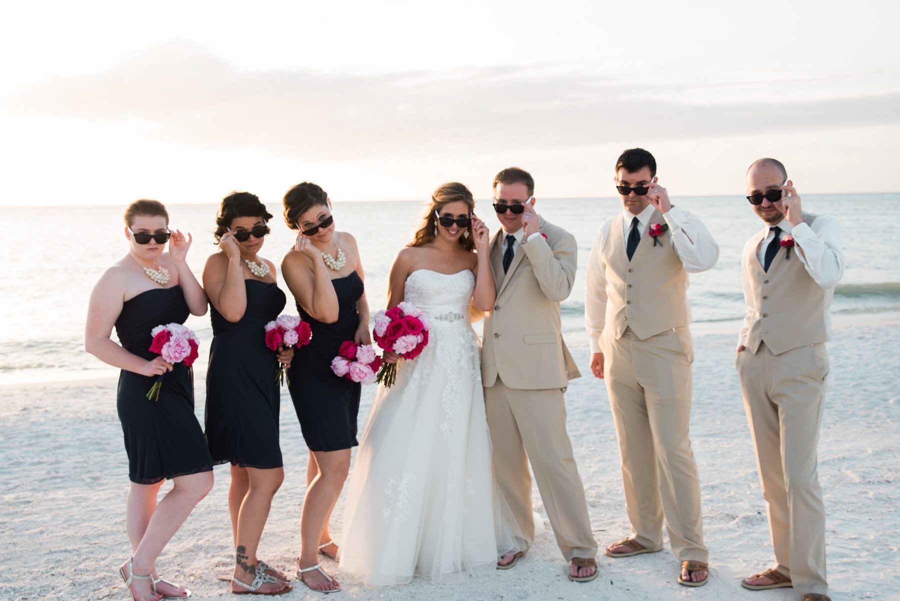 Wedding party on a beach in sunglasses; bride in white gown, bridesmaids in navy, groomsmen in tan.
