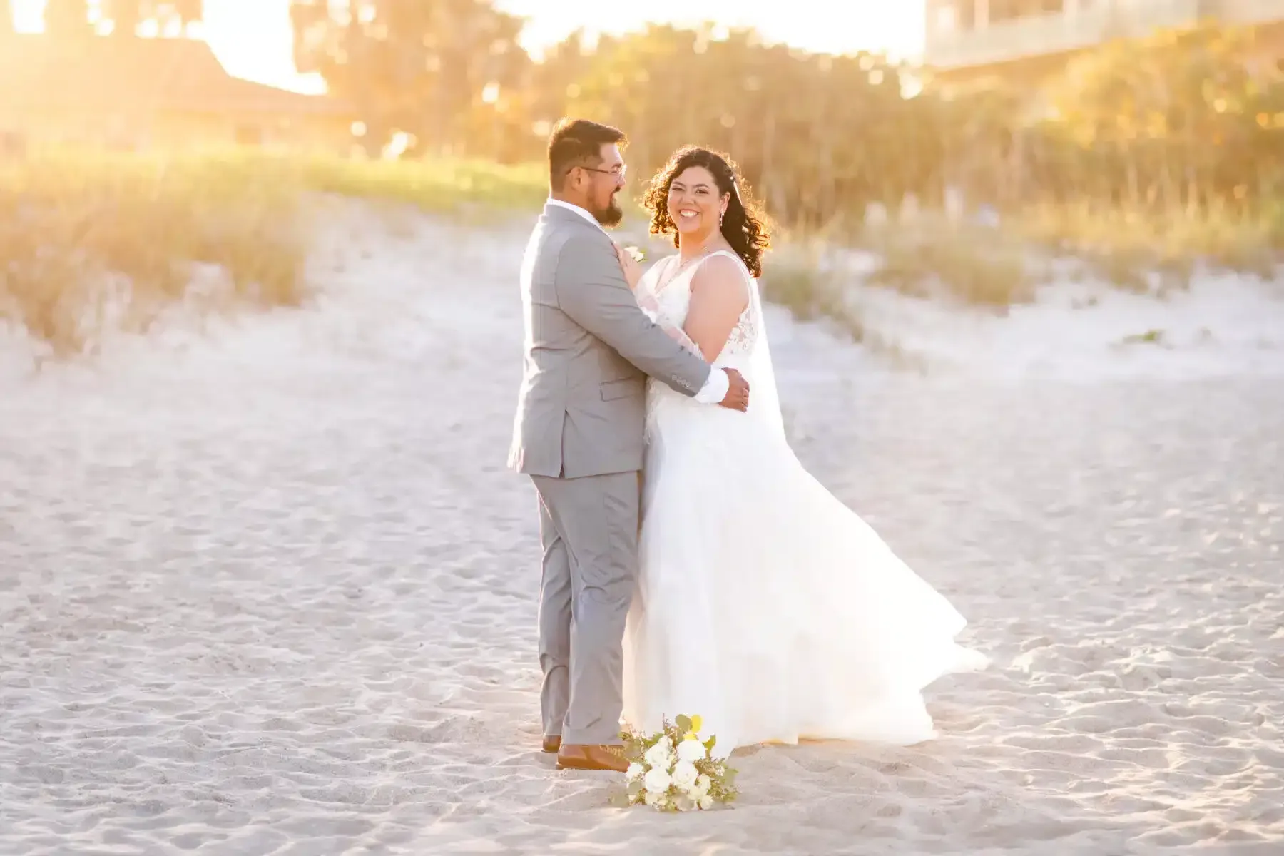 Couple embracing on a beach; woman in wedding dress, man in gray suit. Golden sunlight, flowers nearby.