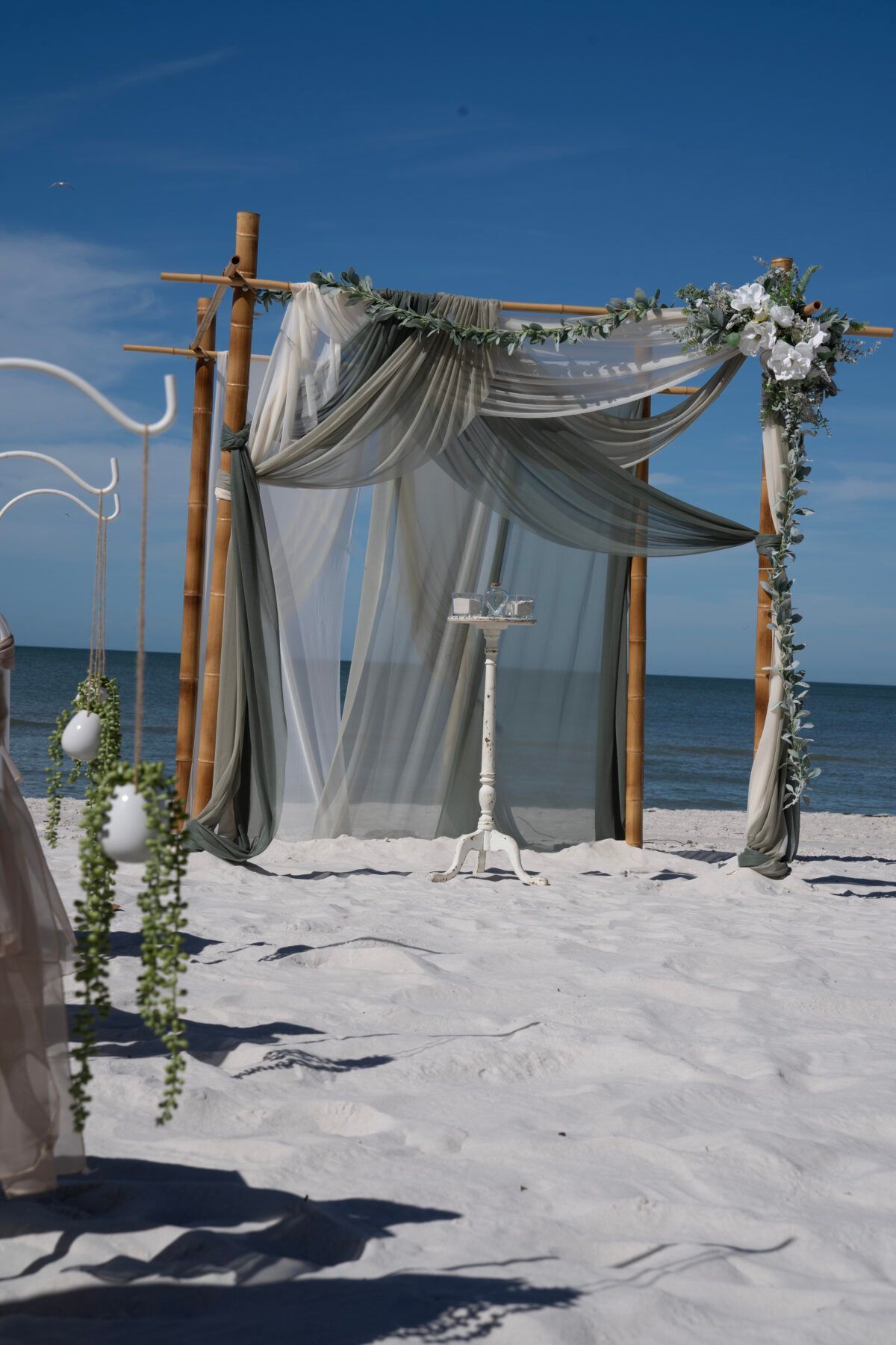 Wedding ceremony setup on a sandy beach under a blue sky, with draped fabric and floral accents.