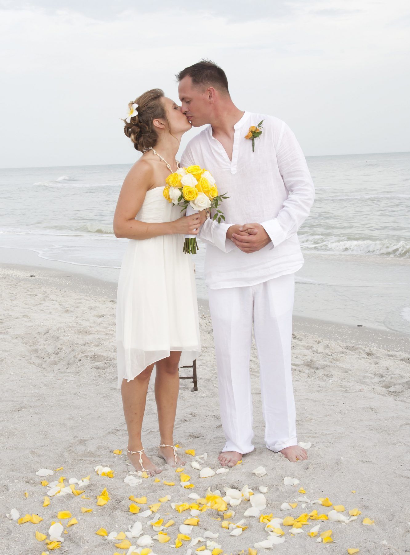 Couple kissing on a beach; bride in white dress holding yellow bouquet, groom in white shirt.