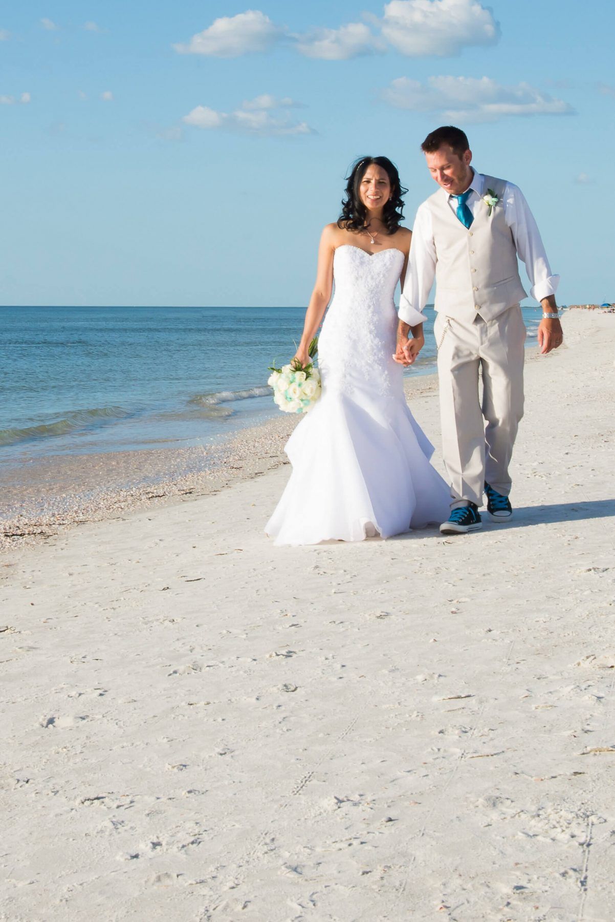 Bride and groom walking hand-in-hand on a sunny beach, holding a bouquet.