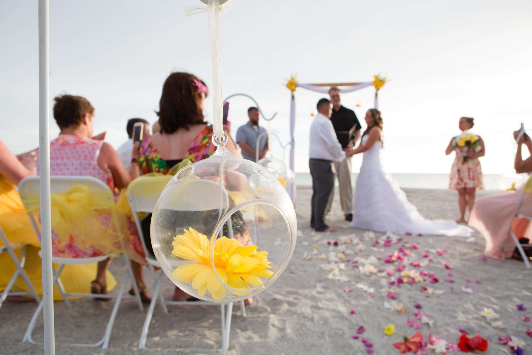 Beach wedding ceremony: bride and groom holding hands under an arch, guests seated, yellow flower decor.