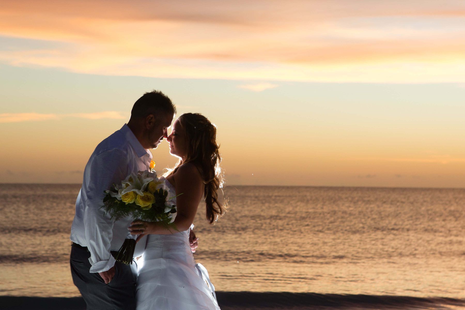 Couple embraces on a beach at sunset; woman in a white dress, man in a white shirt, ocean in the background.